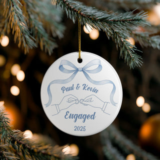 Engagement ornament for gay couple hanging on Christmas tree, showing two men’s hands with rings and blue ribbon accent.