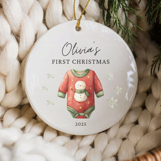 Decorative ornament with 'Olivia's First Christmas 2025' text and baby outfit illustration, placed on a textured surface with greenery.
