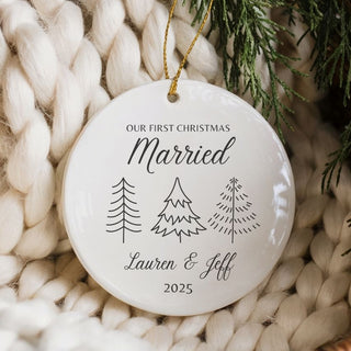 Personalized Christmas ornament with 'Our First Christmas Married' text, trees, and year, placed on a woven surface with white yarn and greenery.