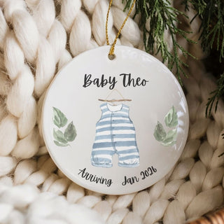 Round ceramic ornament with baby onesie illustration and text on a woven surface