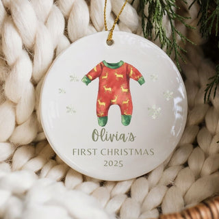 Christmas ornament with a baby onesie design and 'Olivia's First Christmas 2025' text, hanging on a textured surface.