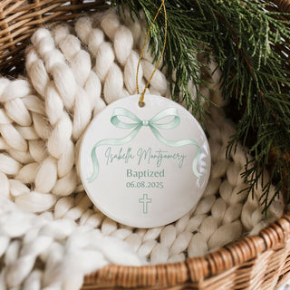 Personalized ornament with a green bow design, text, and a cross, placed on a textured surface with greenery.